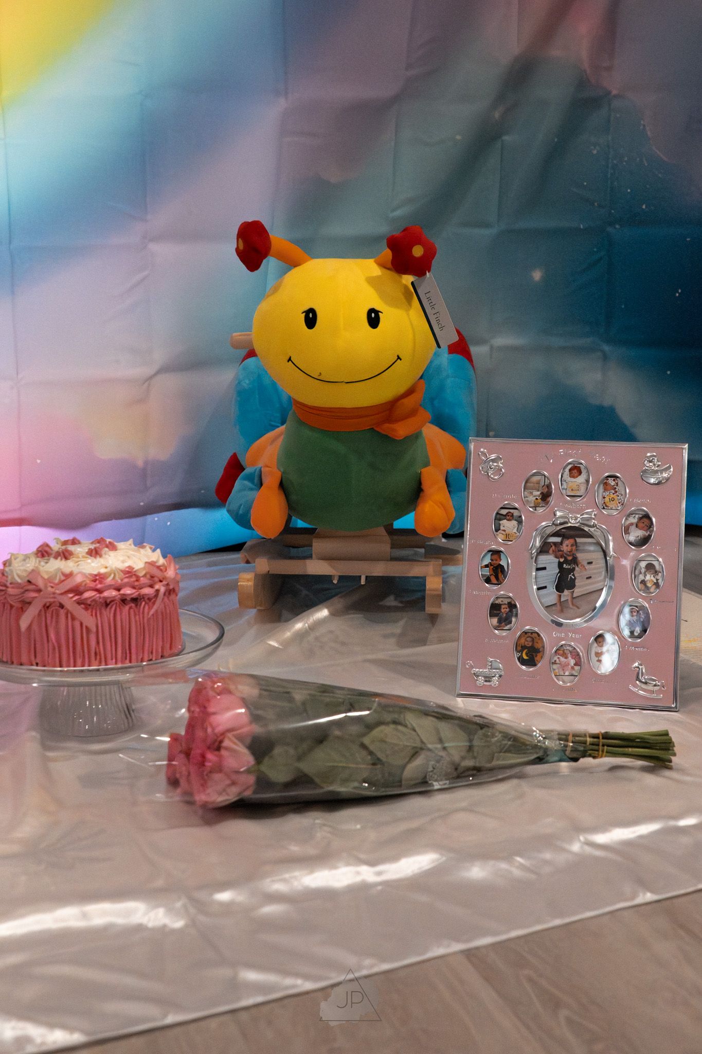 A yellow plush toy sits beside a pink frosted cake, flowers, and a decorative photo frame on a reflective surface.