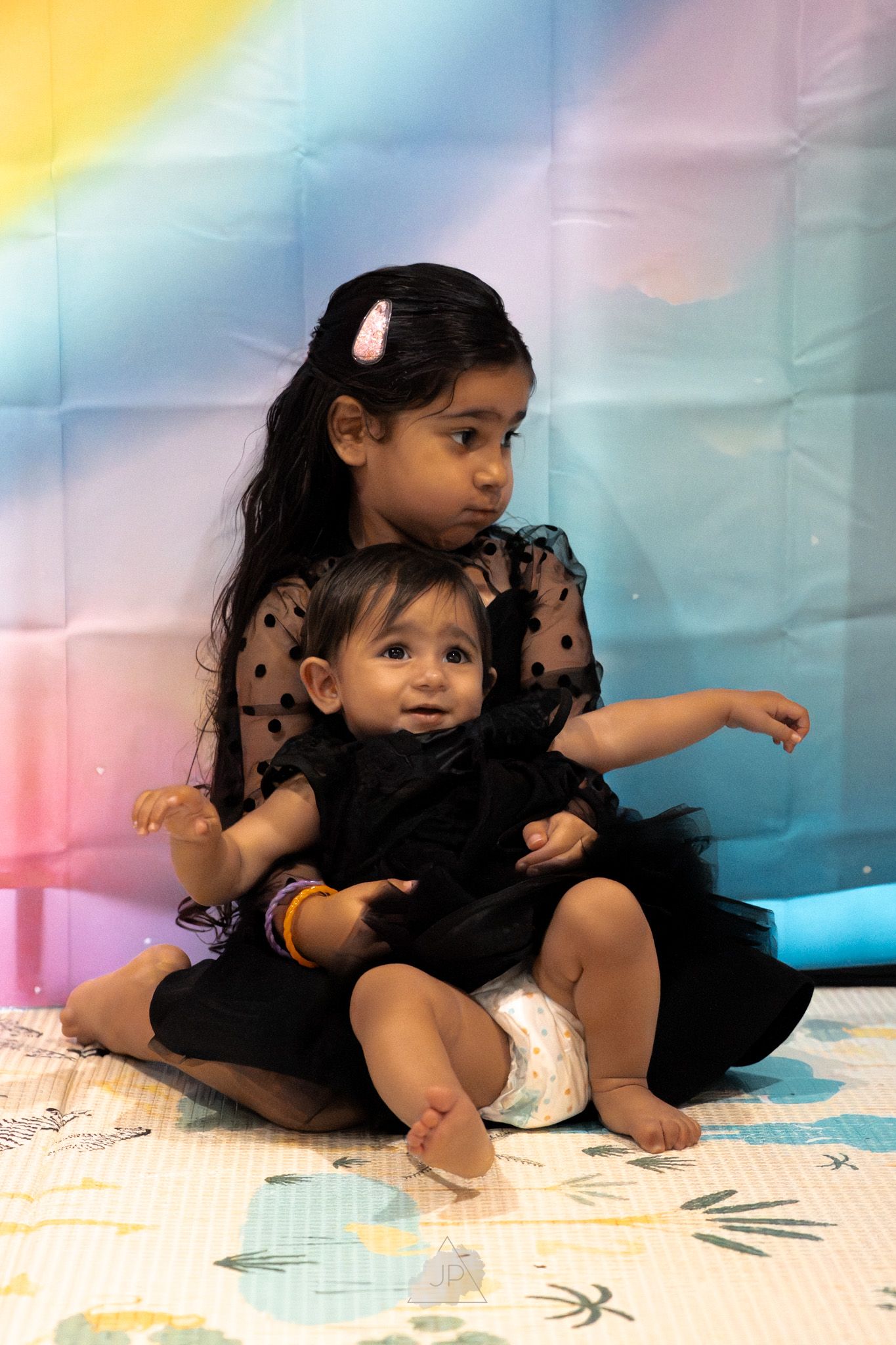 Two children in matching black outfits sit together on a patterned blanket against a pastel rainbow backdrop.