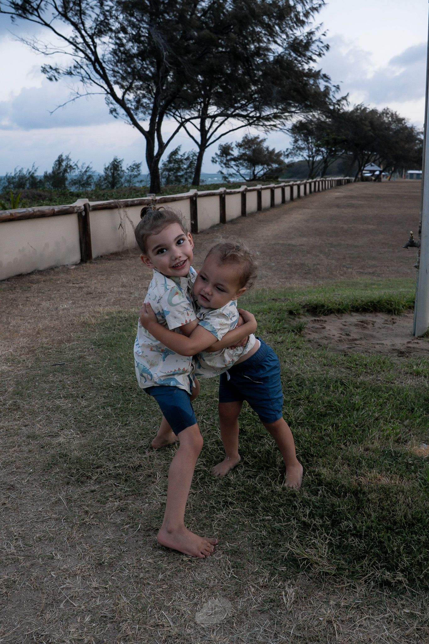 Two children play and hug outdoors on a grassy path lined with trees on a cloudy day.