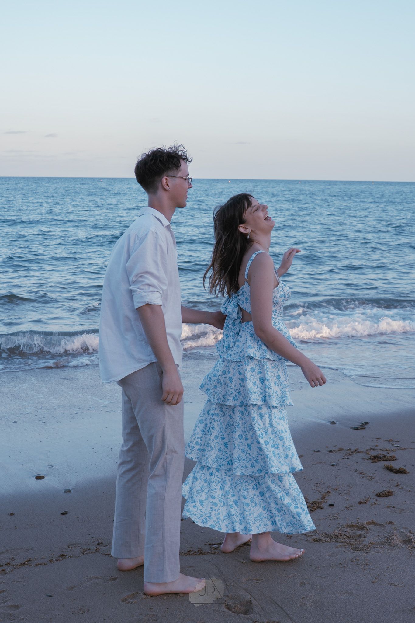 Couple in white and blue outfits dancing carefree on a sandy beach with waves rolling in.