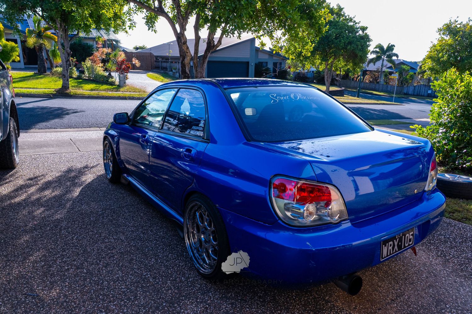 Blue Subaru WRX with large rear spoiler parked on driveway with trees visible in background.