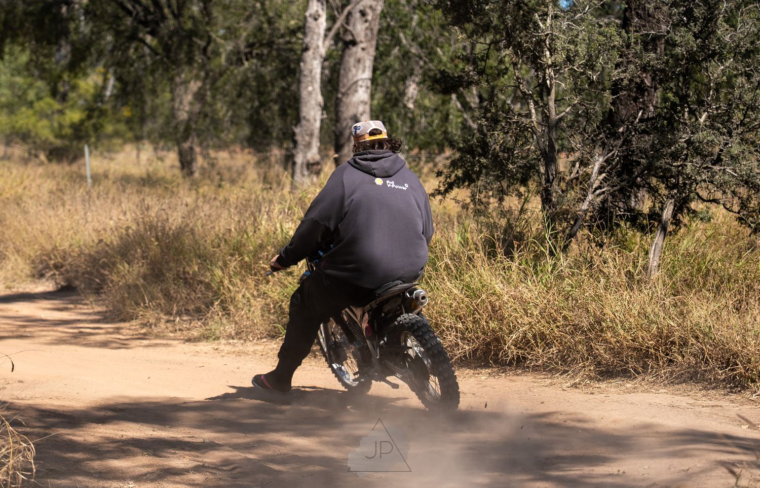 A motorcycle rider navigates a dusty dirt road surrounded by trees and brush in a rural setting.