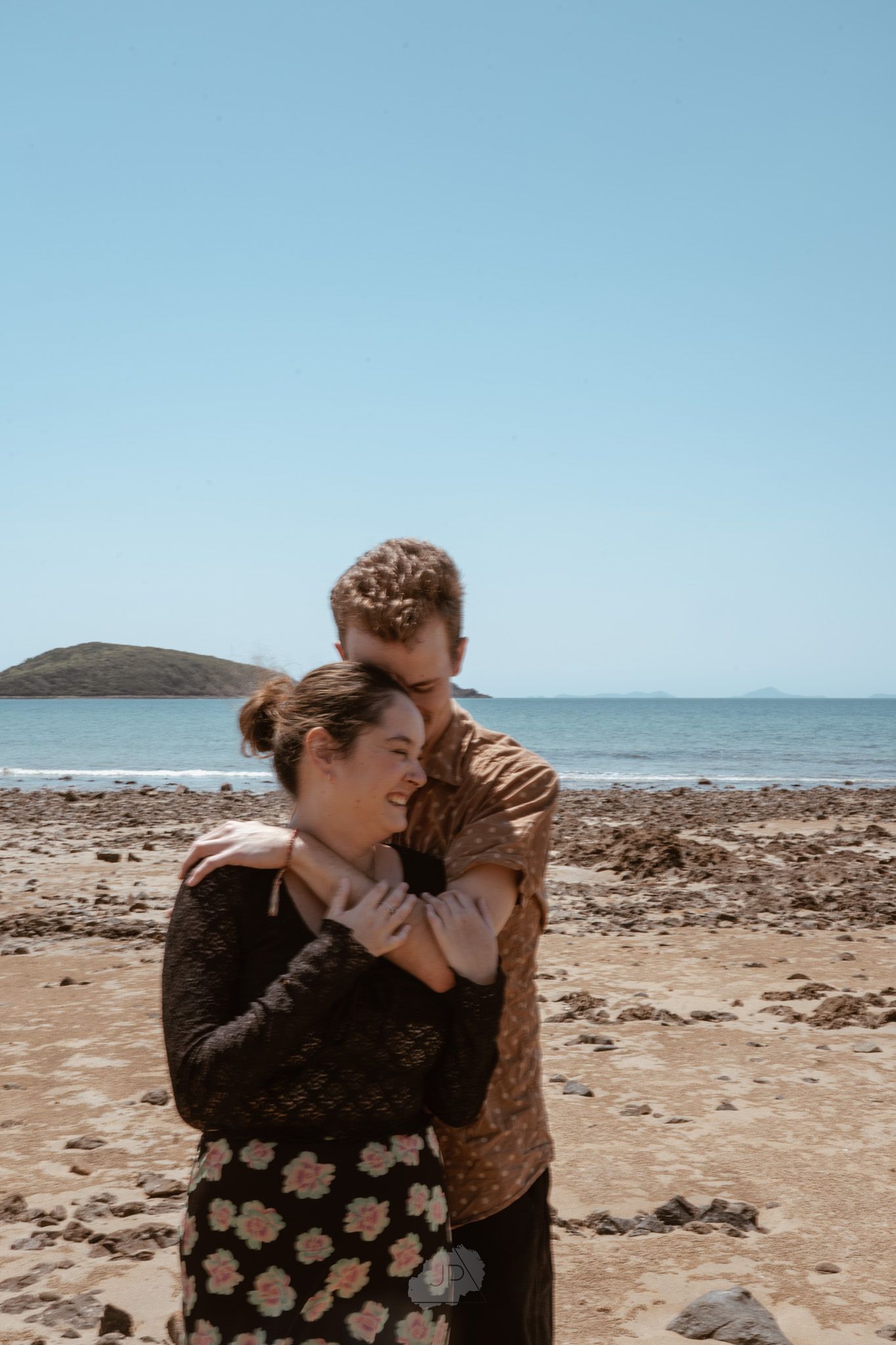 A couple shares a romantic moment on a sunny beach with rocky terrain and ocean views.