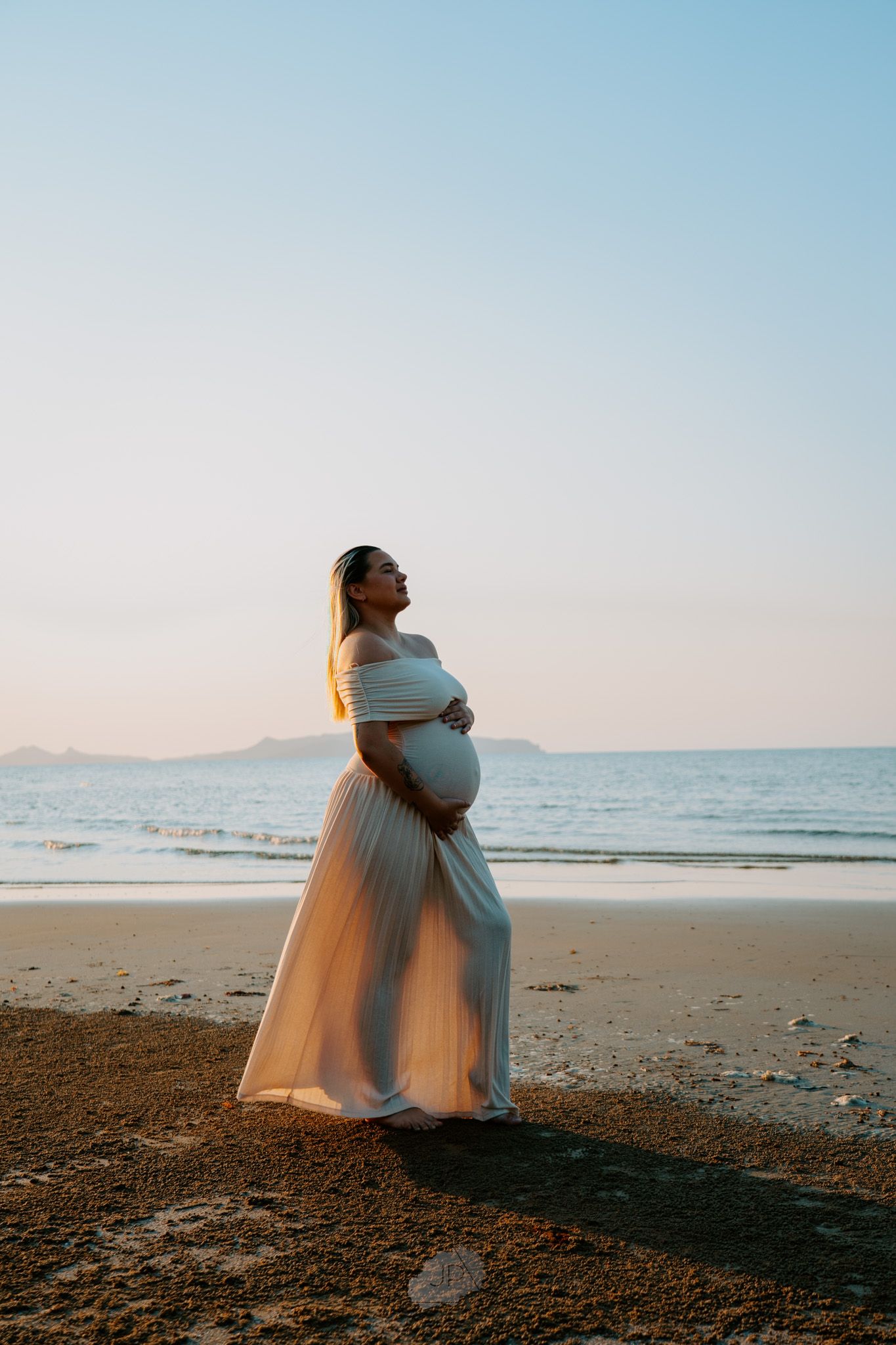 A silhouetted figure in a flowing white dress stands on a beach at sunset, cradling their pregnant belly.