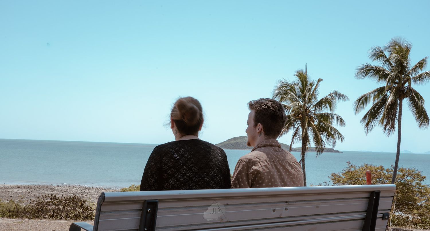 A couple enjoys a romantic sunset view from a bench overlooking the ocean with palm trees swaying in the breeze.