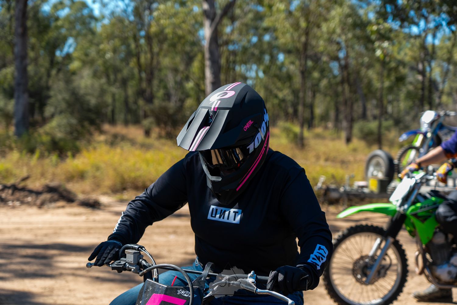 A rider in black protective gear on a dirt bike navigates a trail with another motorcycle visible in the background.