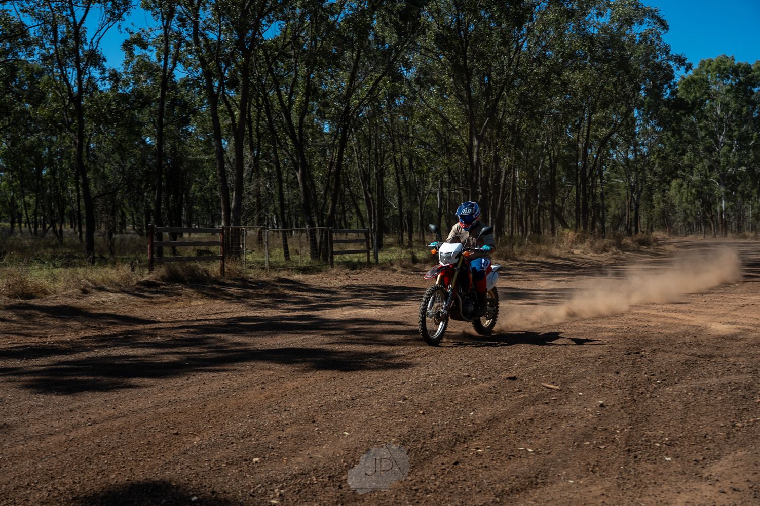 A person rides a dirt bike along a dusty trail through Australian bushland on a sunny day.