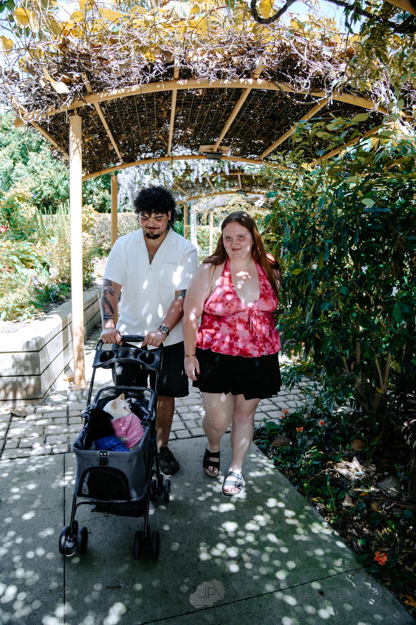 Two people relax together on a bench under a shaded garden pergola on a sunny afternoon.