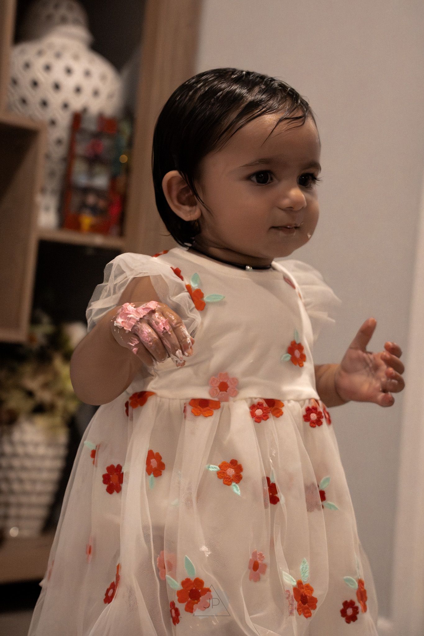 A small child wearing a white dress with orange floral embellishments stands indoors in warm lighting.