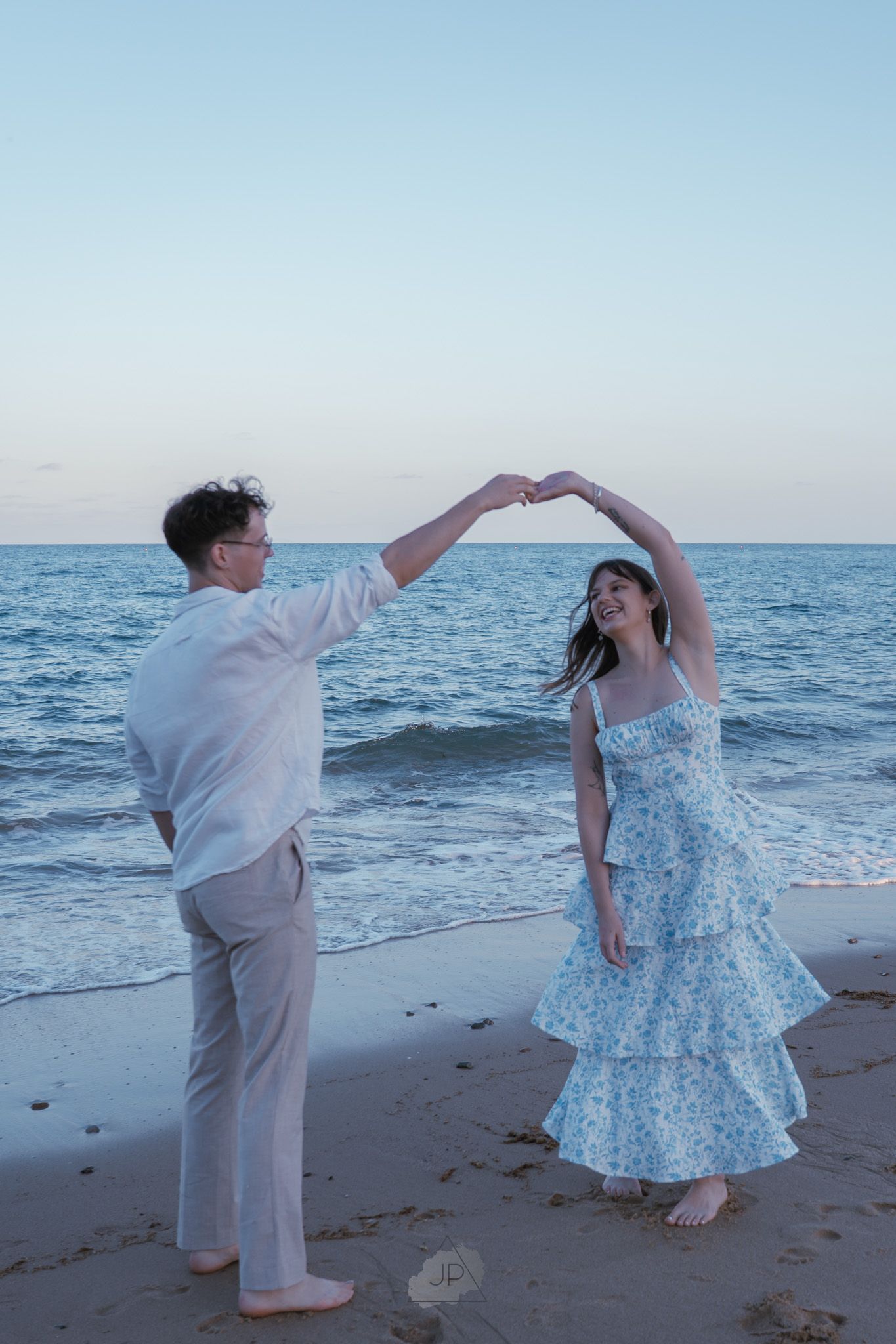 A couple in light colored outfits dance together on a beach at sunset with the ocean waves in the background.
