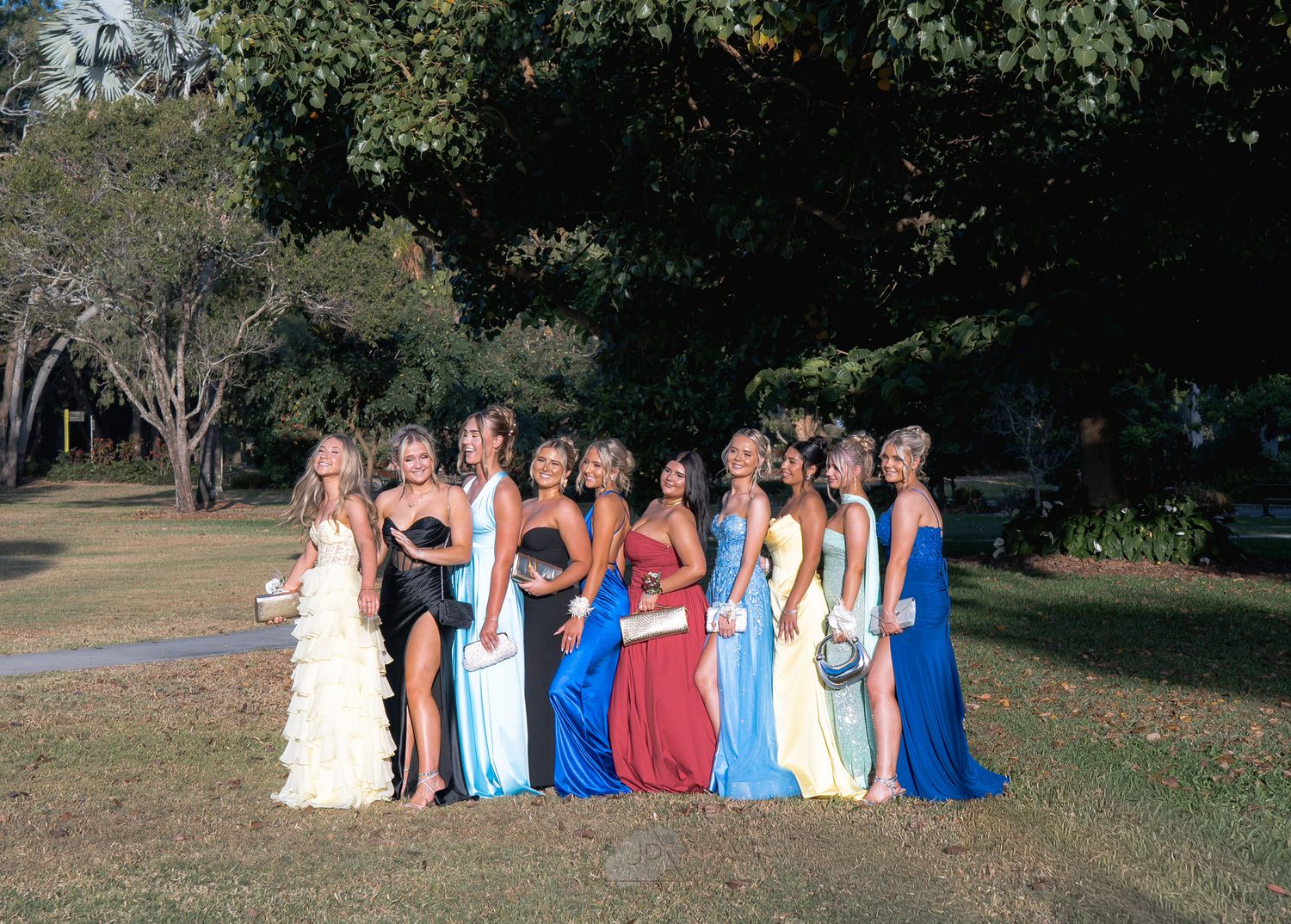 A group in formal attire poses together outdoors on a sunny day for a special occasion photo.