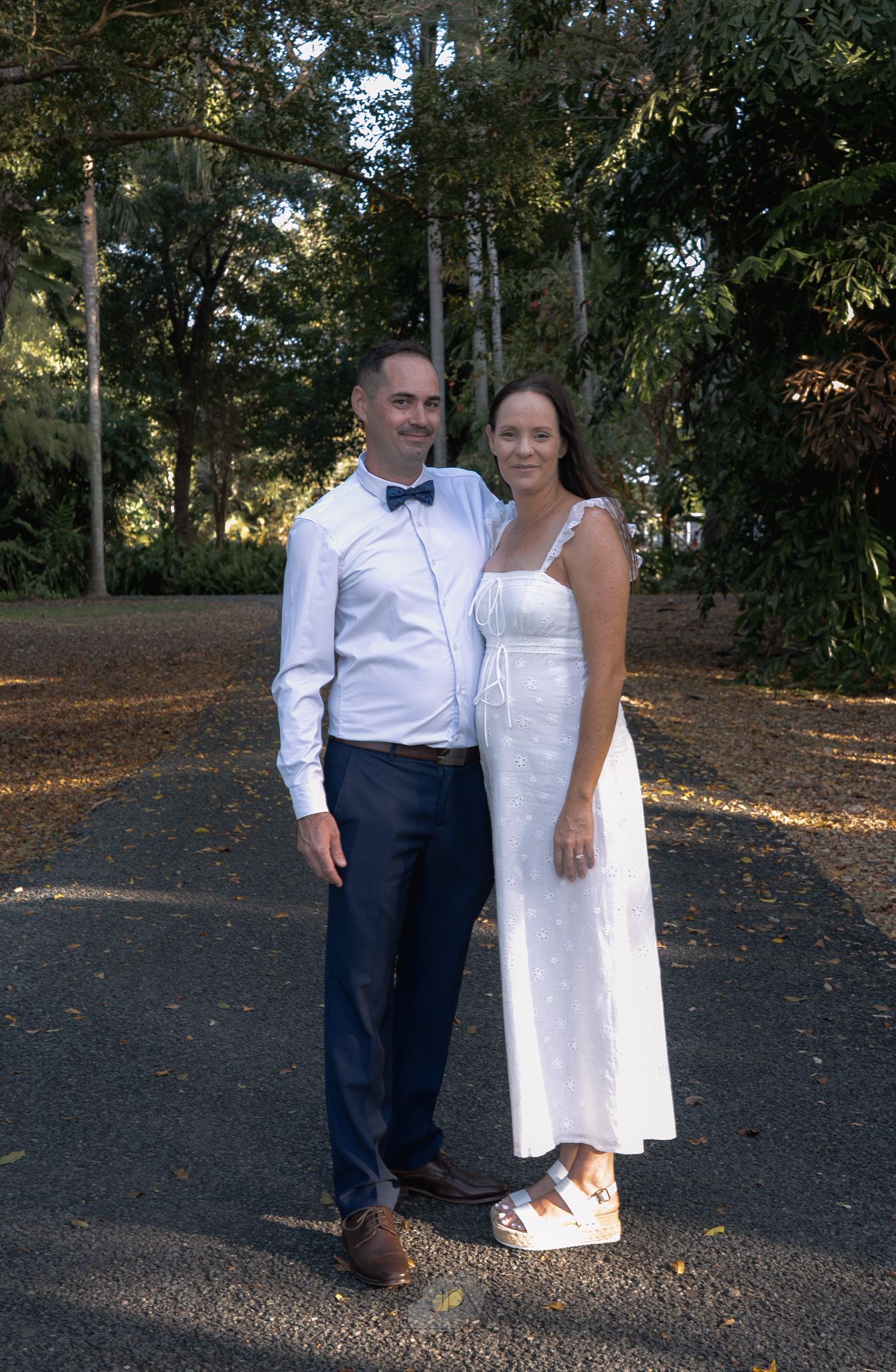 A couple in white and navy attire poses together outdoors on a wooded path during golden hour.
