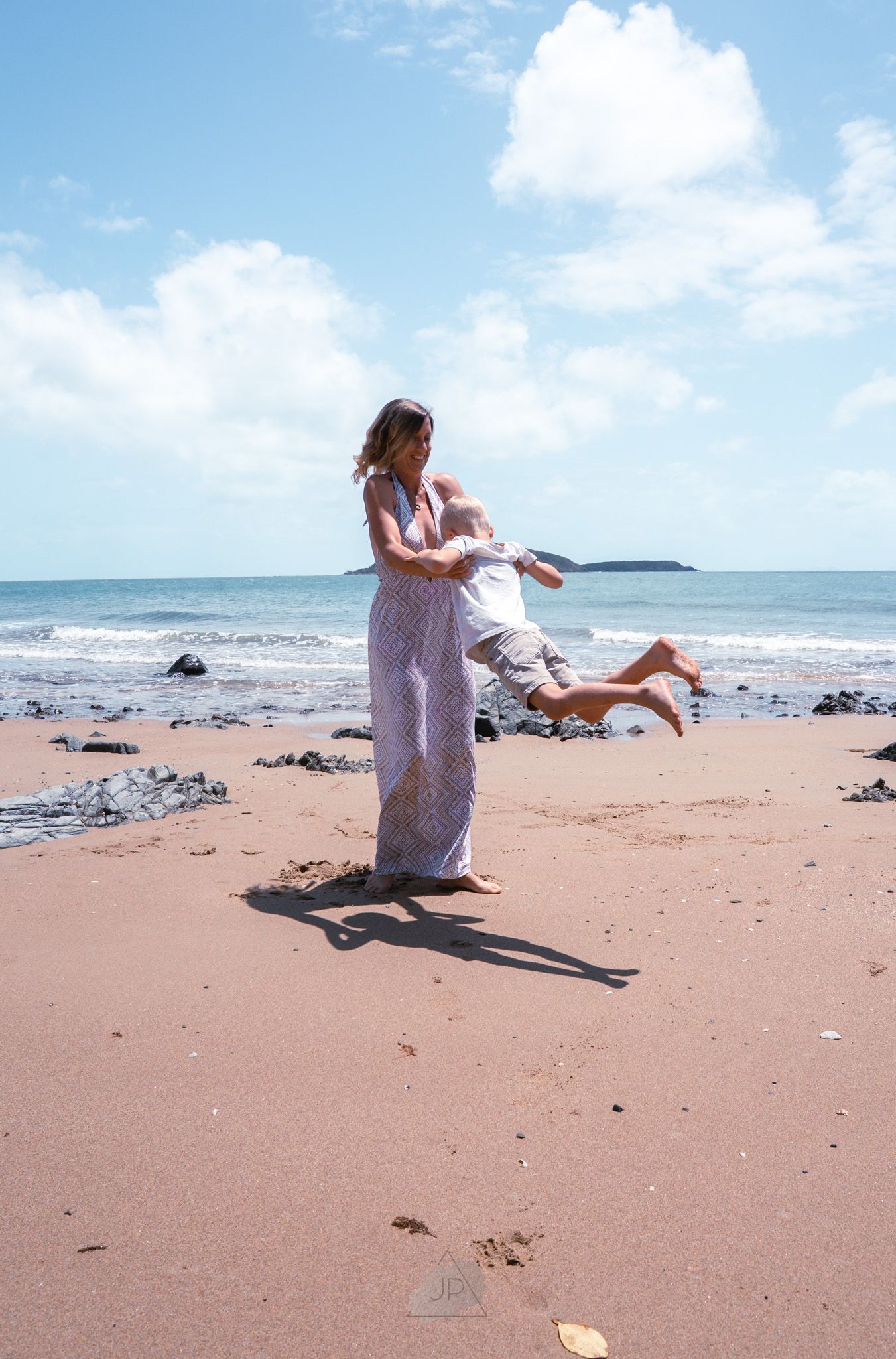 A person in flowy pants spins a small child playfully on a scenic beach with blue sky and ocean in the background.