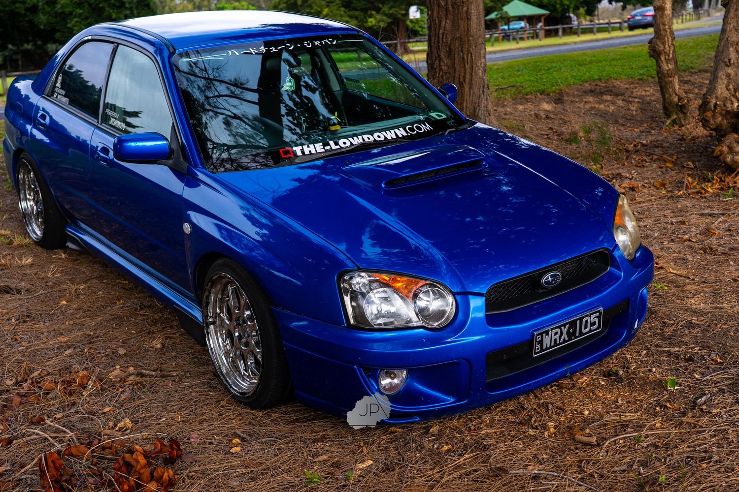 Bright blue modified Subaru Impreza WRX parked on shaded pavement with custom wheels and body kit.