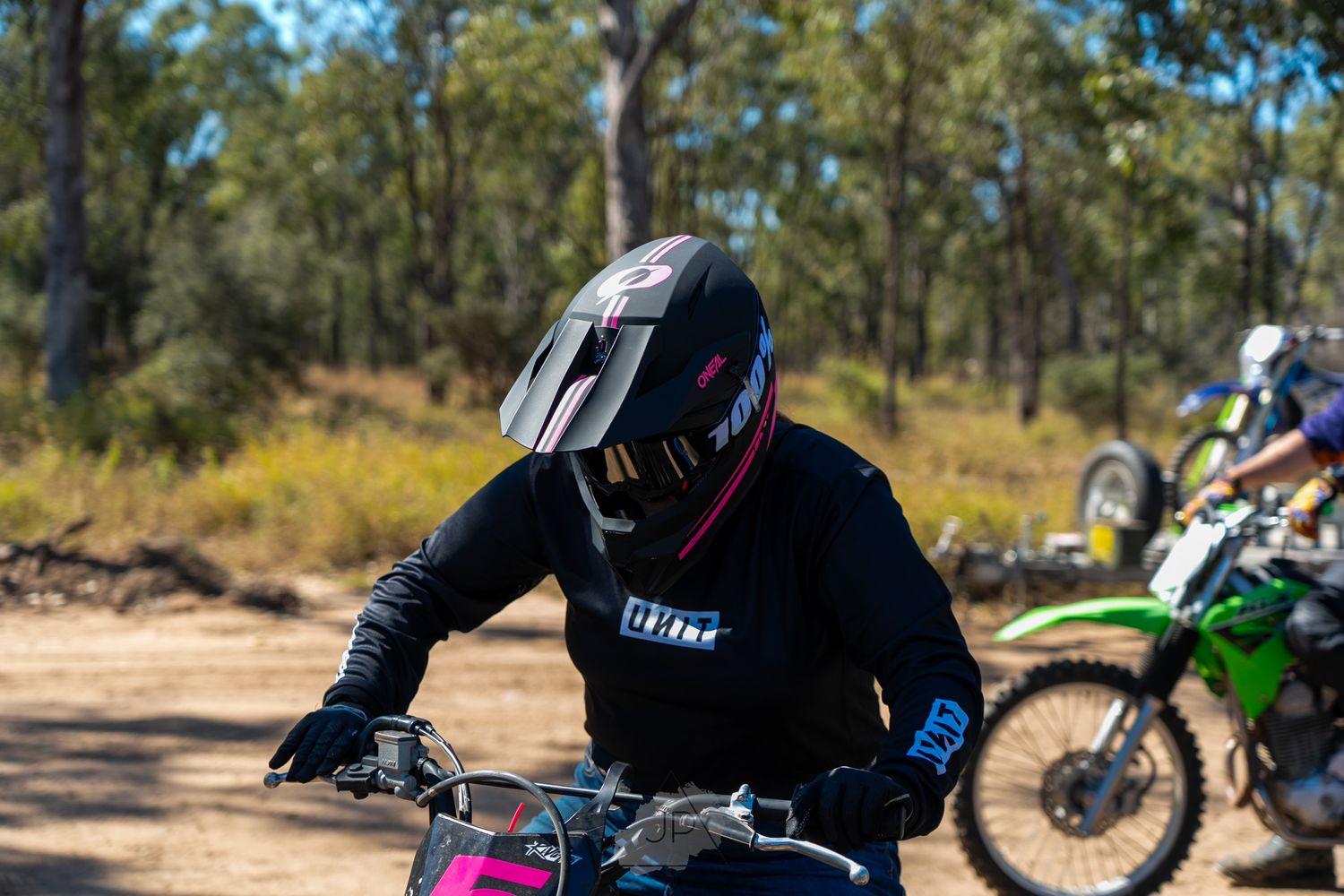 Dirt bike riders gather on a dusty off-road trail surrounded by trees on a sunny day.