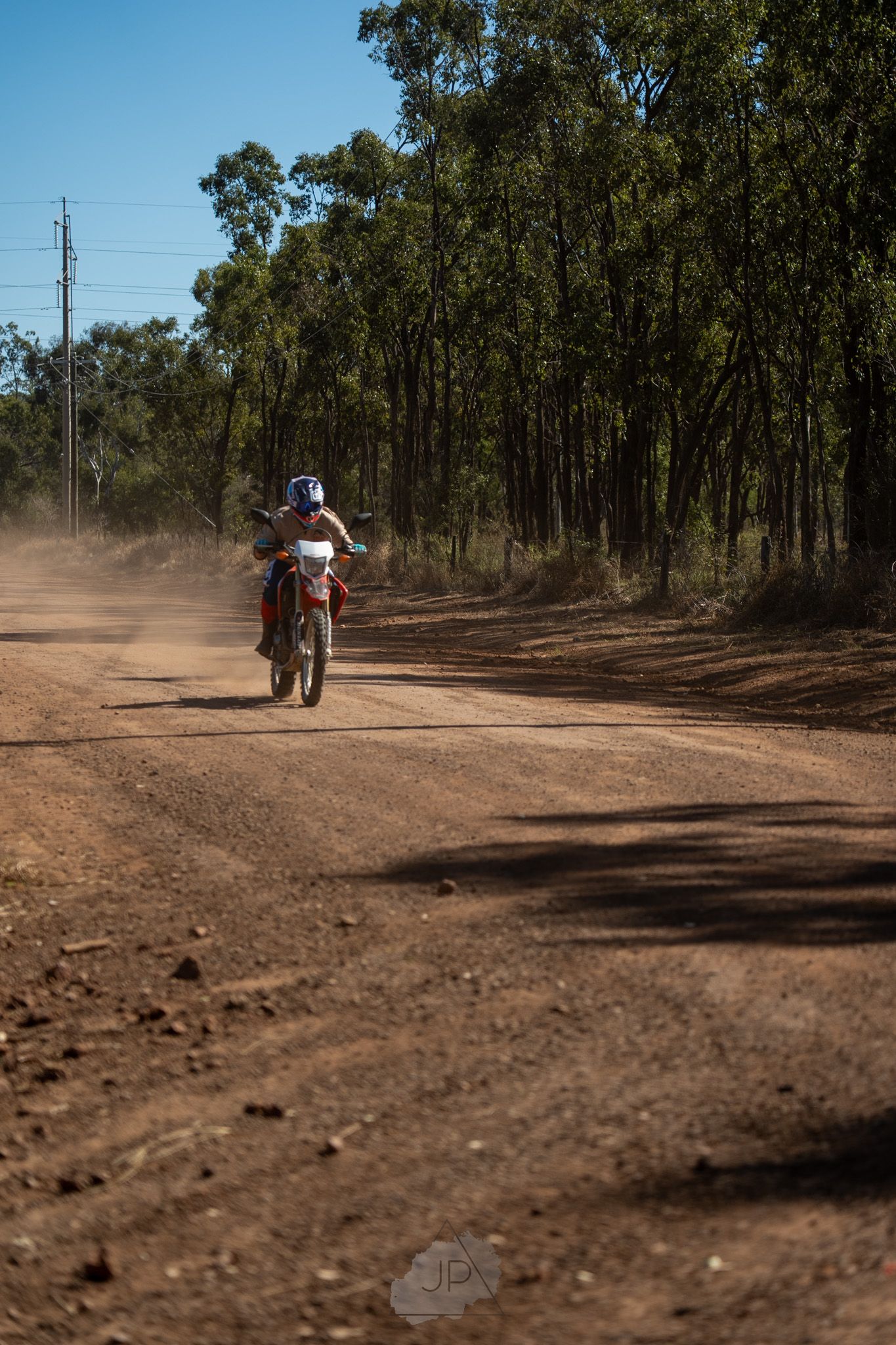 Dirt path curving through Australian bush with motorcycles kicking up dust on a dry terrain.
