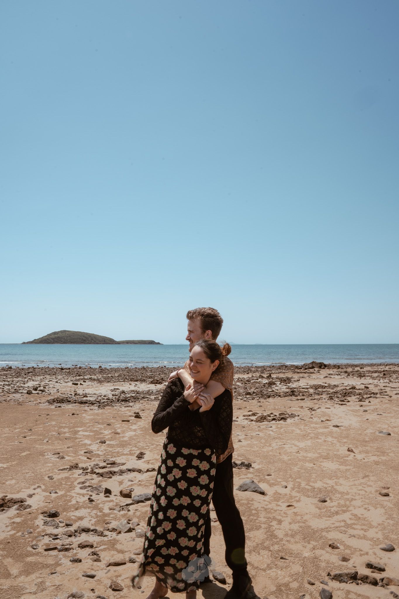 Couple embraces on a rocky beach with ocean and distant island views on a sunny day.