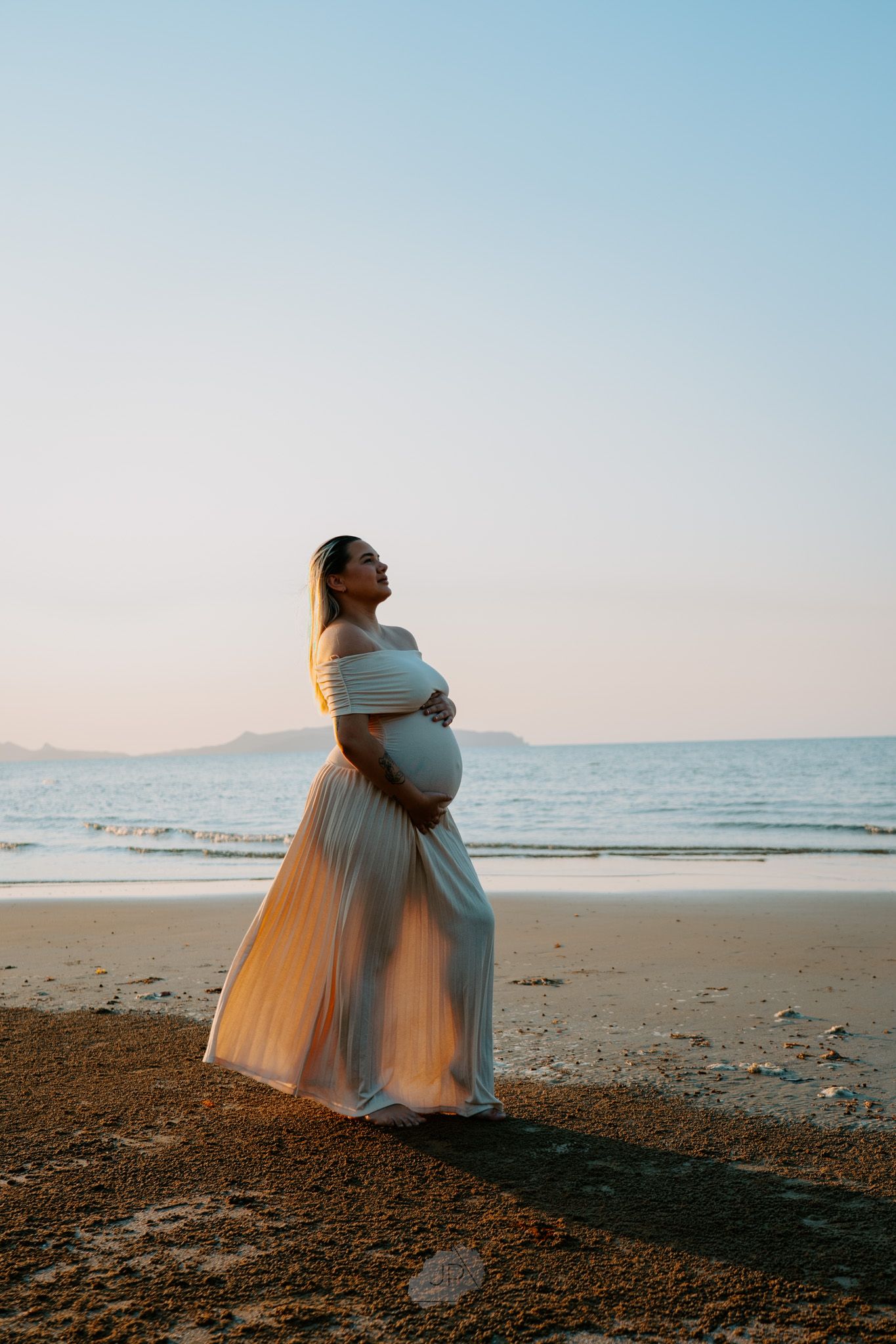 A person in a flowing white dress stands on a sandy beach at sunset, creating a romantic silhouette against the ocean.