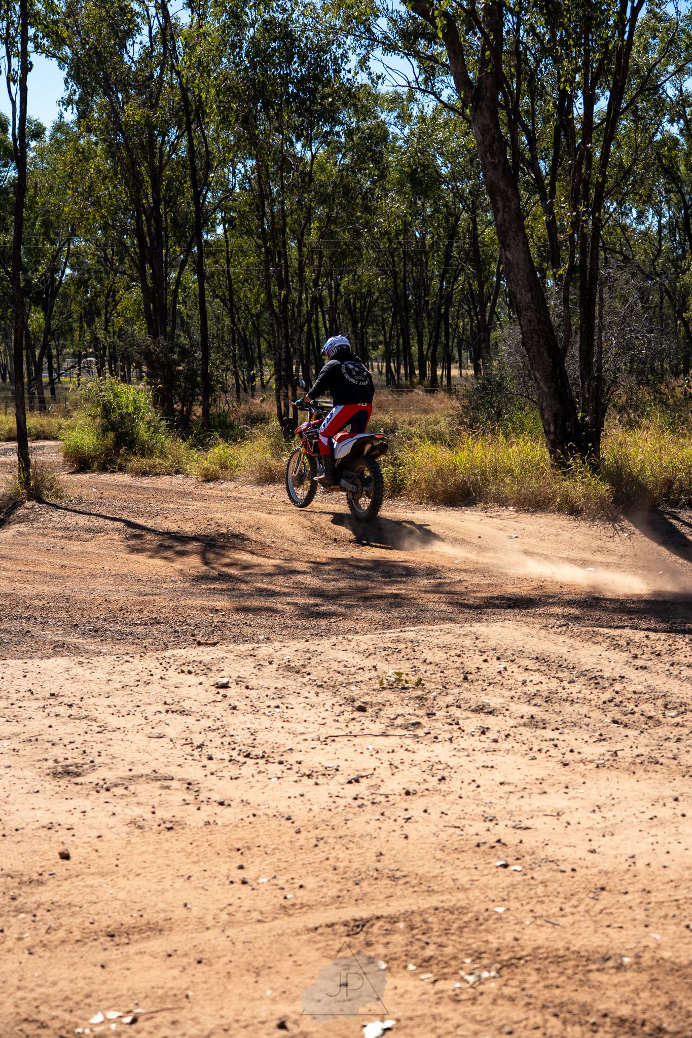 A dirt bike rider navigates a sandy off-road trail through eucalyptus trees on a sunny day.