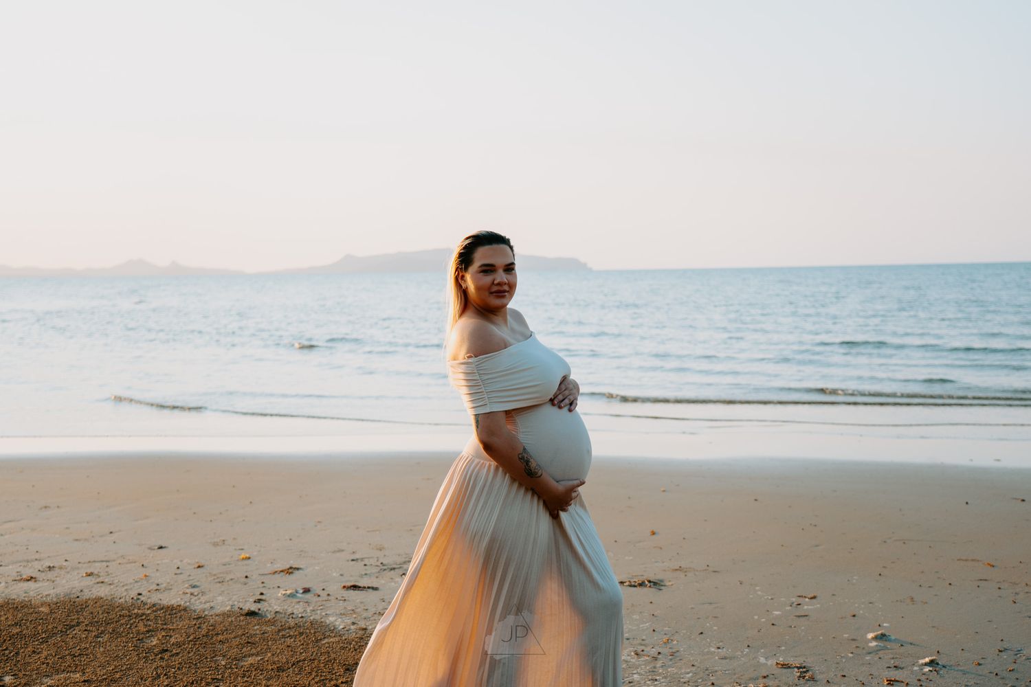 Silhouette of expectant mother in white dress standing on sandy beach during golden hour.