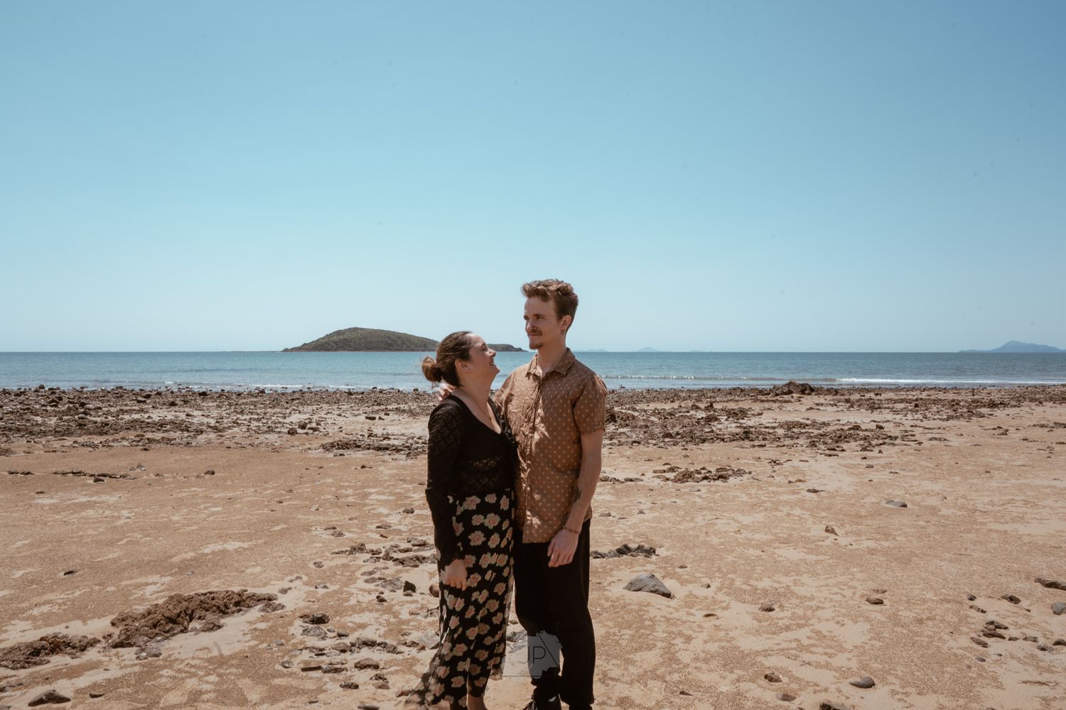 A couple walks along a scenic beach shoreline with mountains visible in the distance on a sunny day.