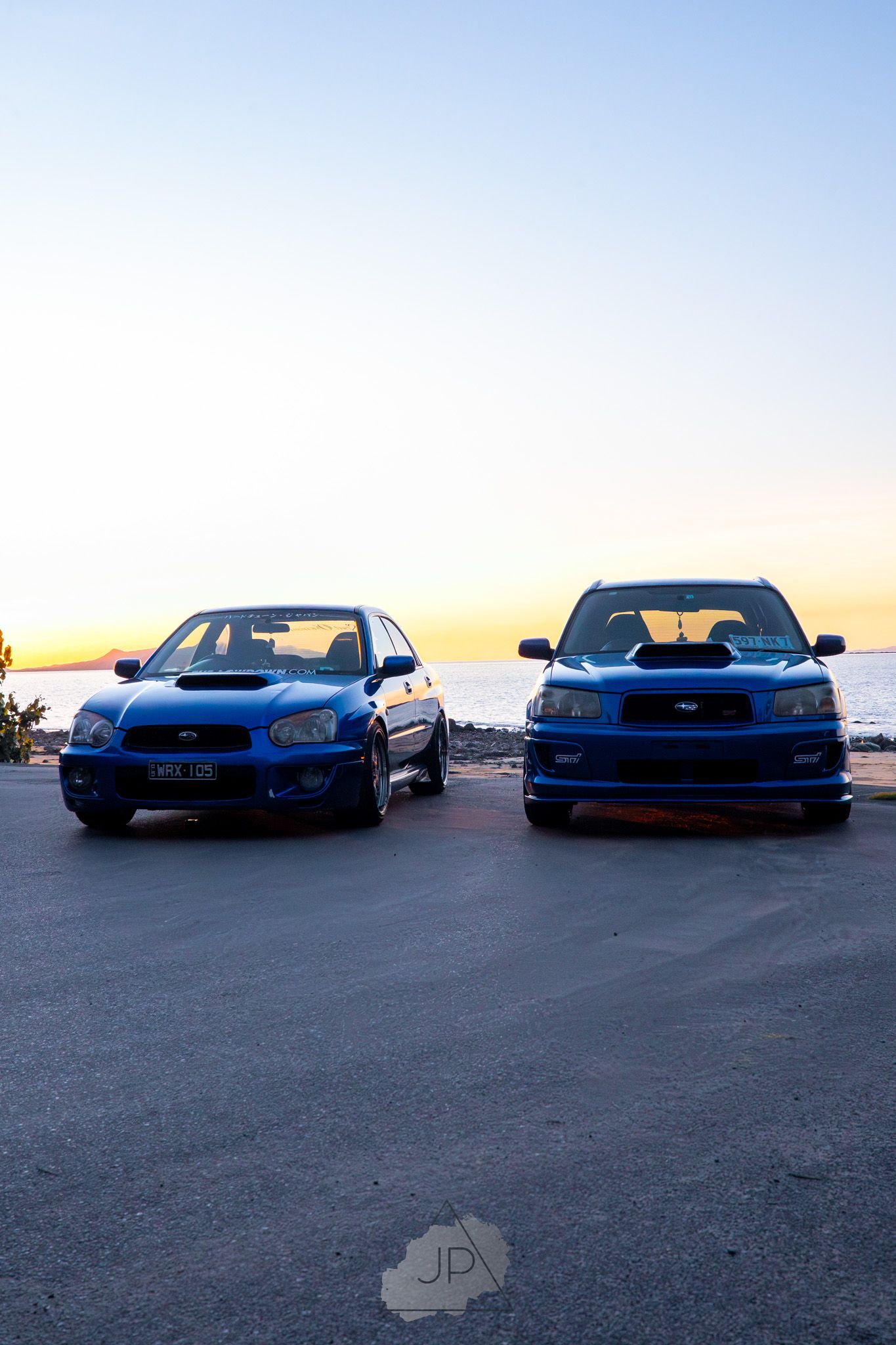 Two Subaru WRX STI cars parked side by side against a sunset sky background.