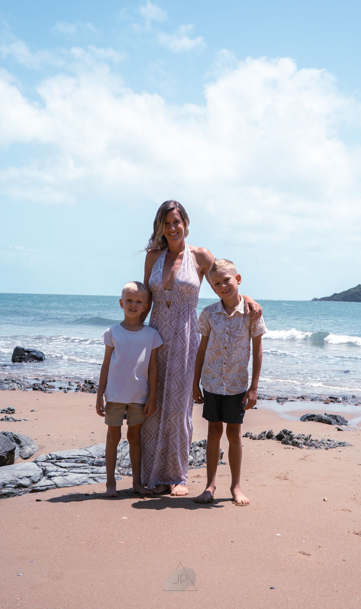 Three people stand together on a sandy beach with ocean waves and rocky coastline in the background.