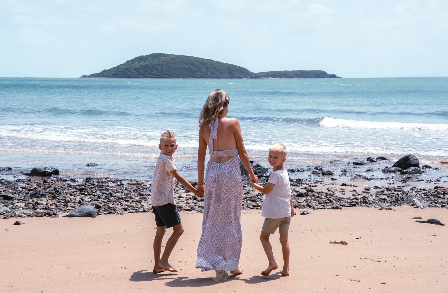 A family walks along the shoreline of a scenic beach with mountains visible in the distance.