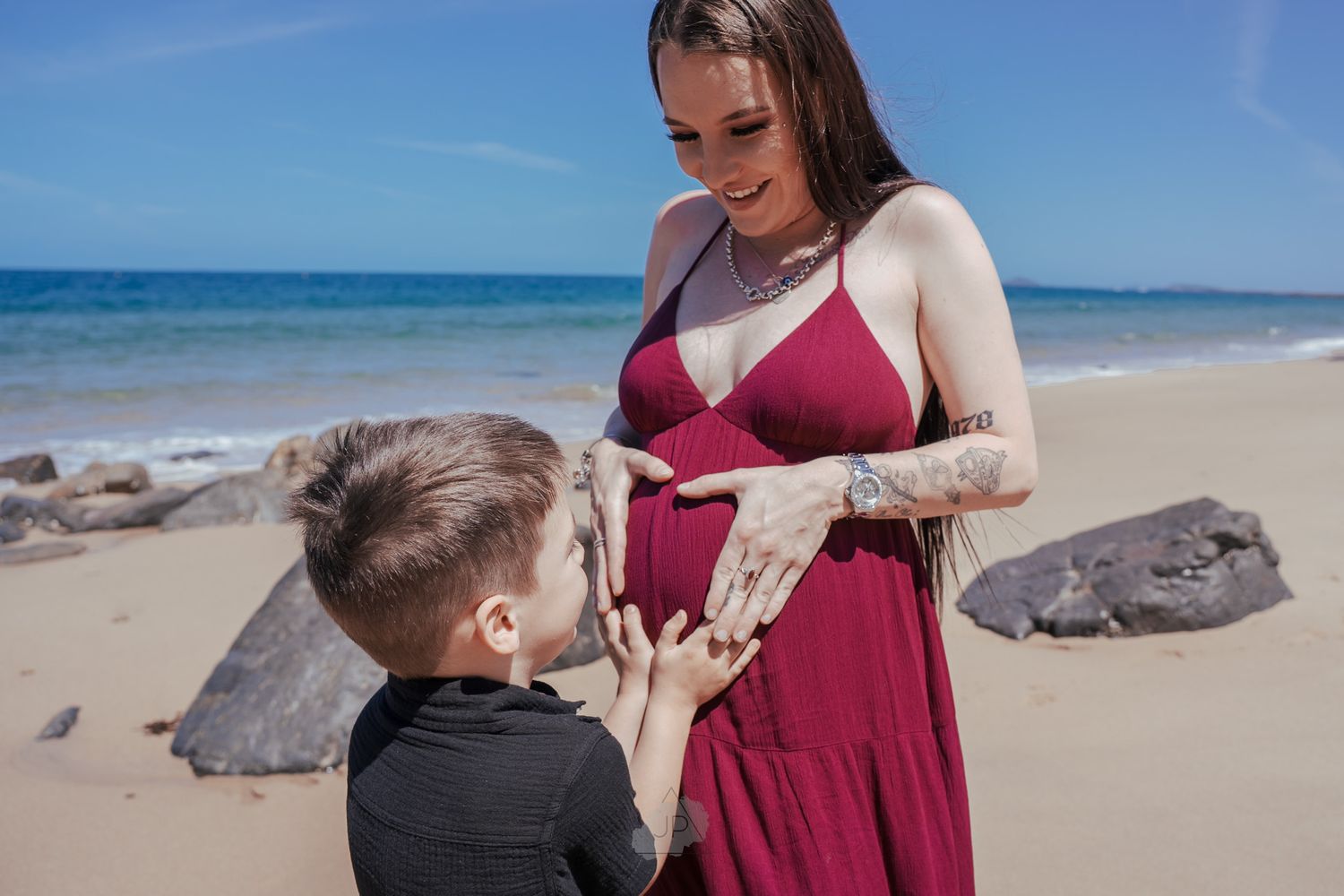 A touching moment on a sunny beach as a child in black reaches toward a pregnant person in a burgundy dress.