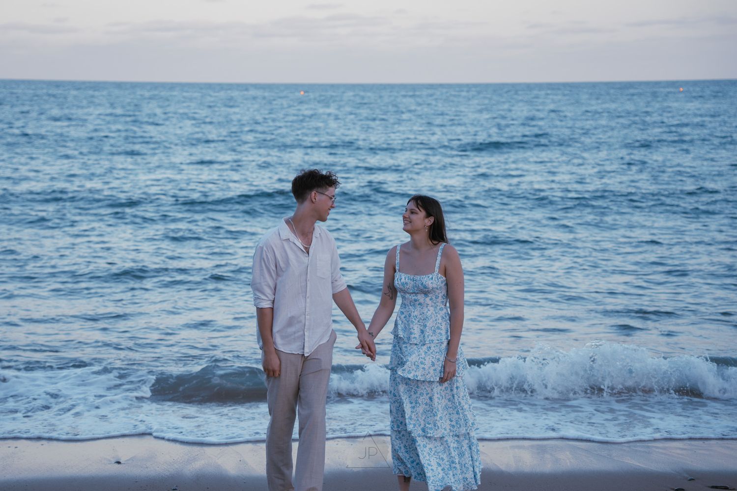 Couple in white outfits walking along a beach during sunset while waves crash on the shore.