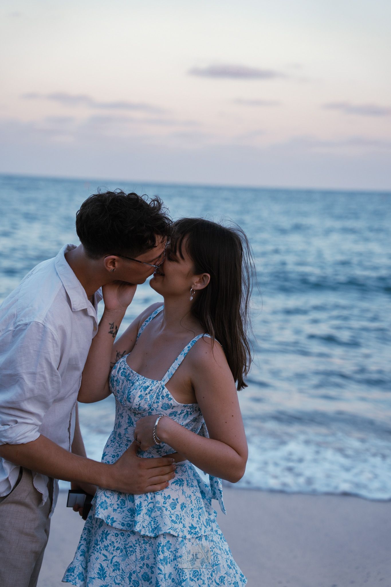 A couple shares a romantic moment on the beach at sunset with ocean waves in the background.