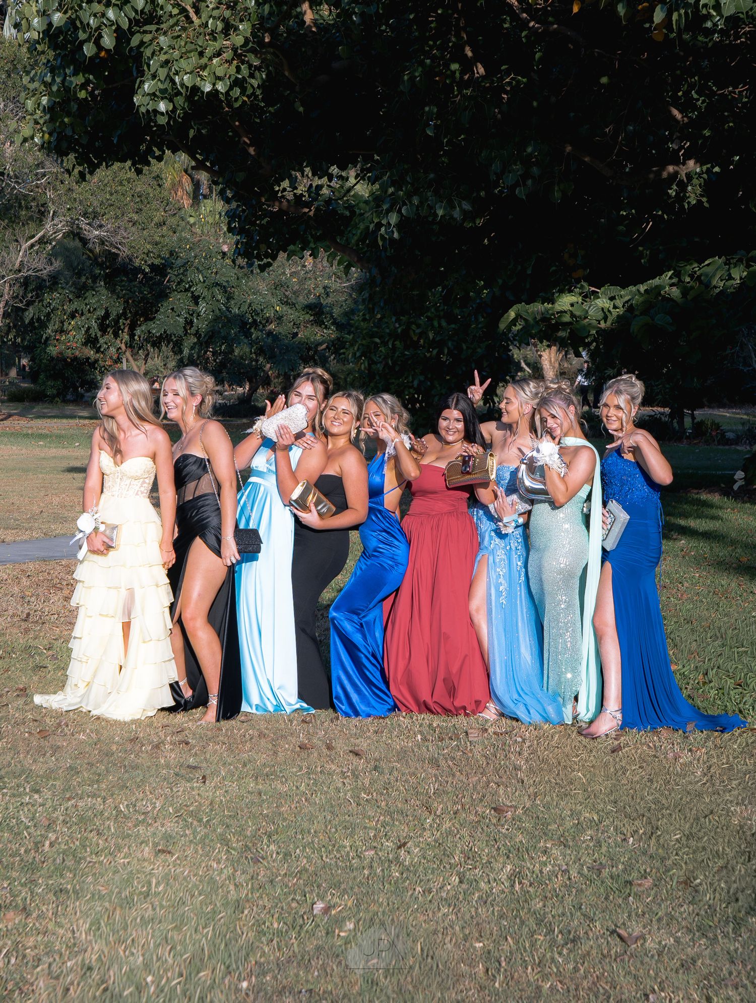 A group in coordinated blue and coral dresses sits in a row on grassy ground during an outdoor photo session.