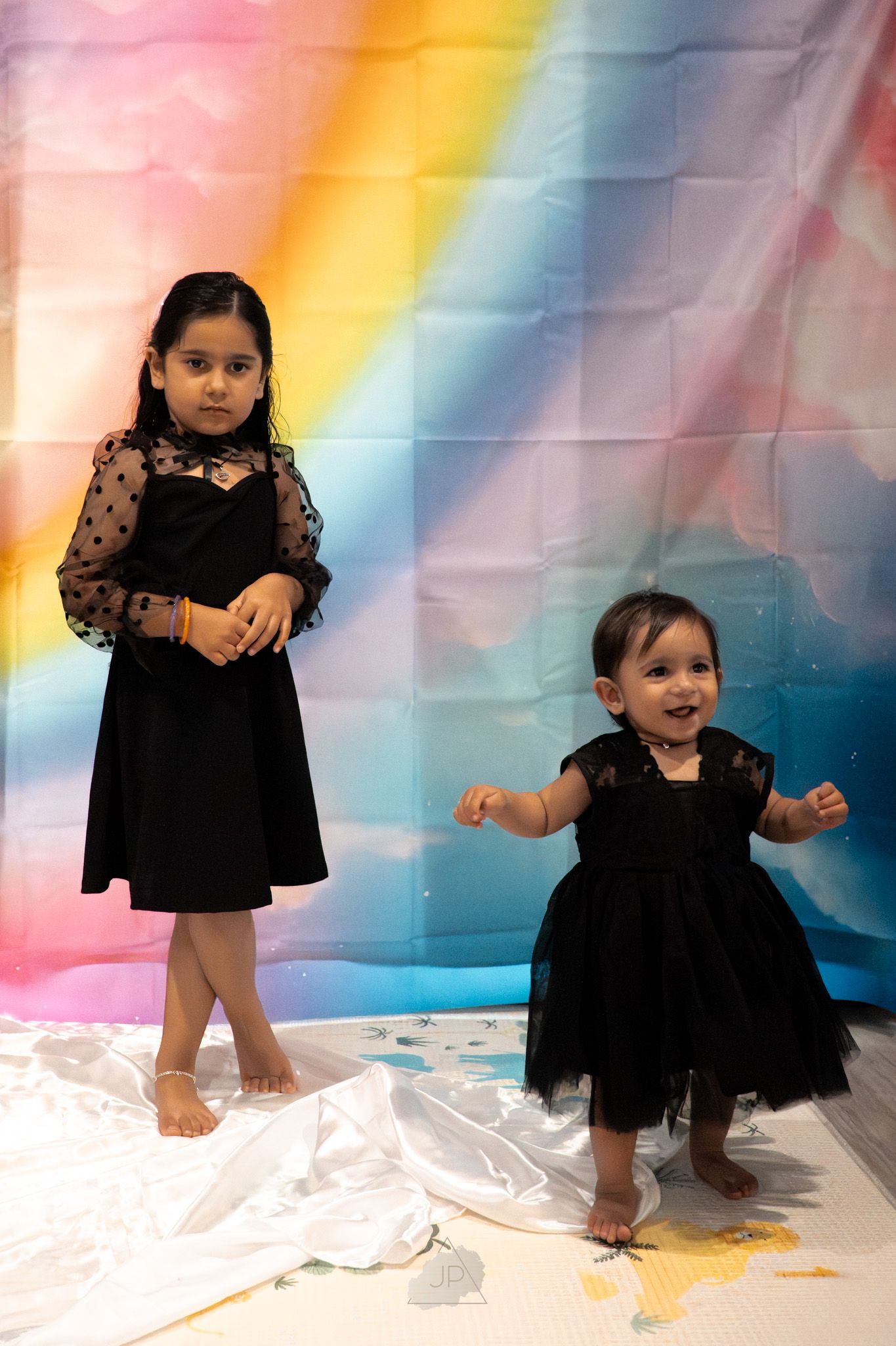 Two children in black lace dresses pose against a colorful rainbow backdrop with white fabric on the floor.