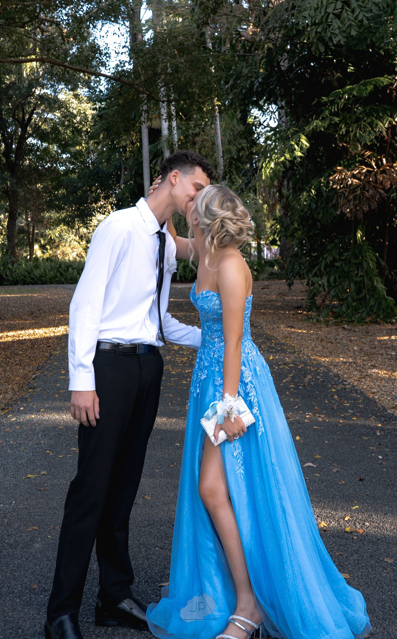 A couple in formal attire share a romantic moment outdoors, with her wearing a flowing blue gown with a slit.