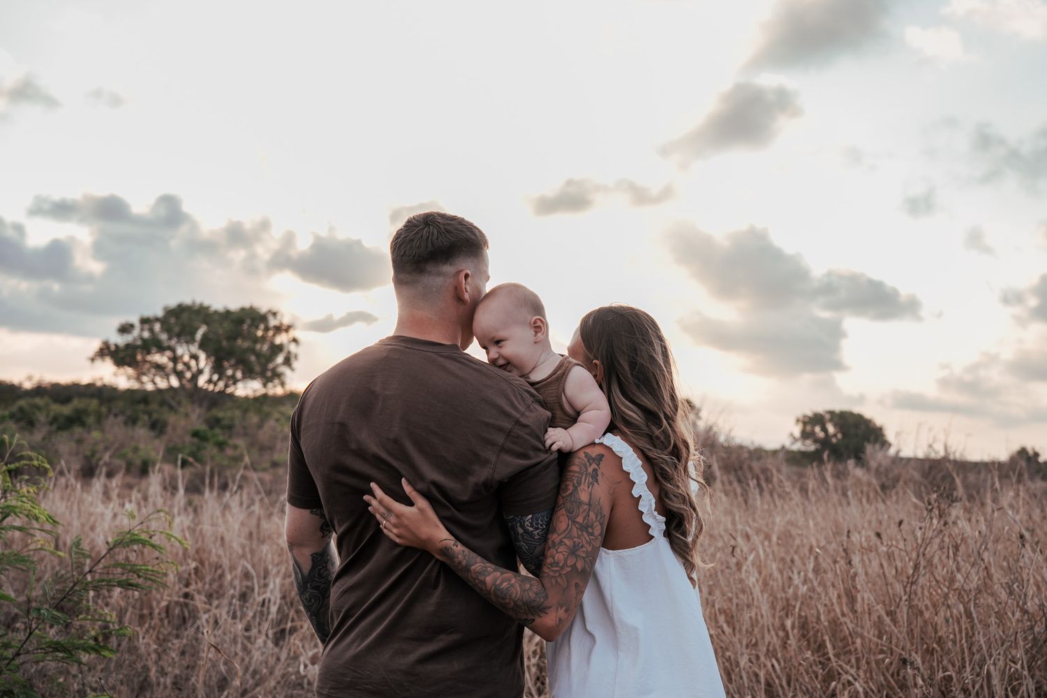 A family embraces in a field of tall grass during golden hour as the sun sets behind them.