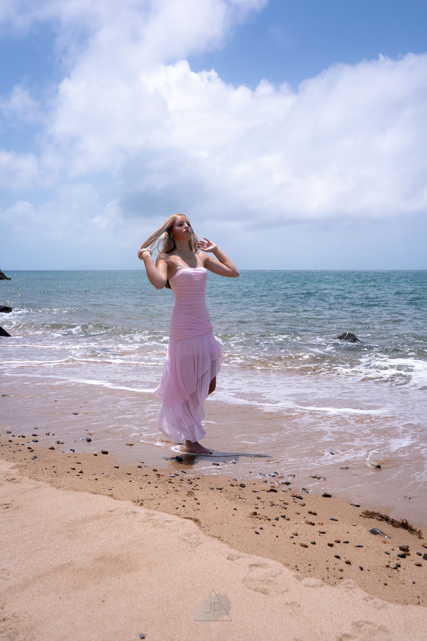 A person in a flowing pink dress twirls gracefully on a sunny beach with waves crashing in the background.