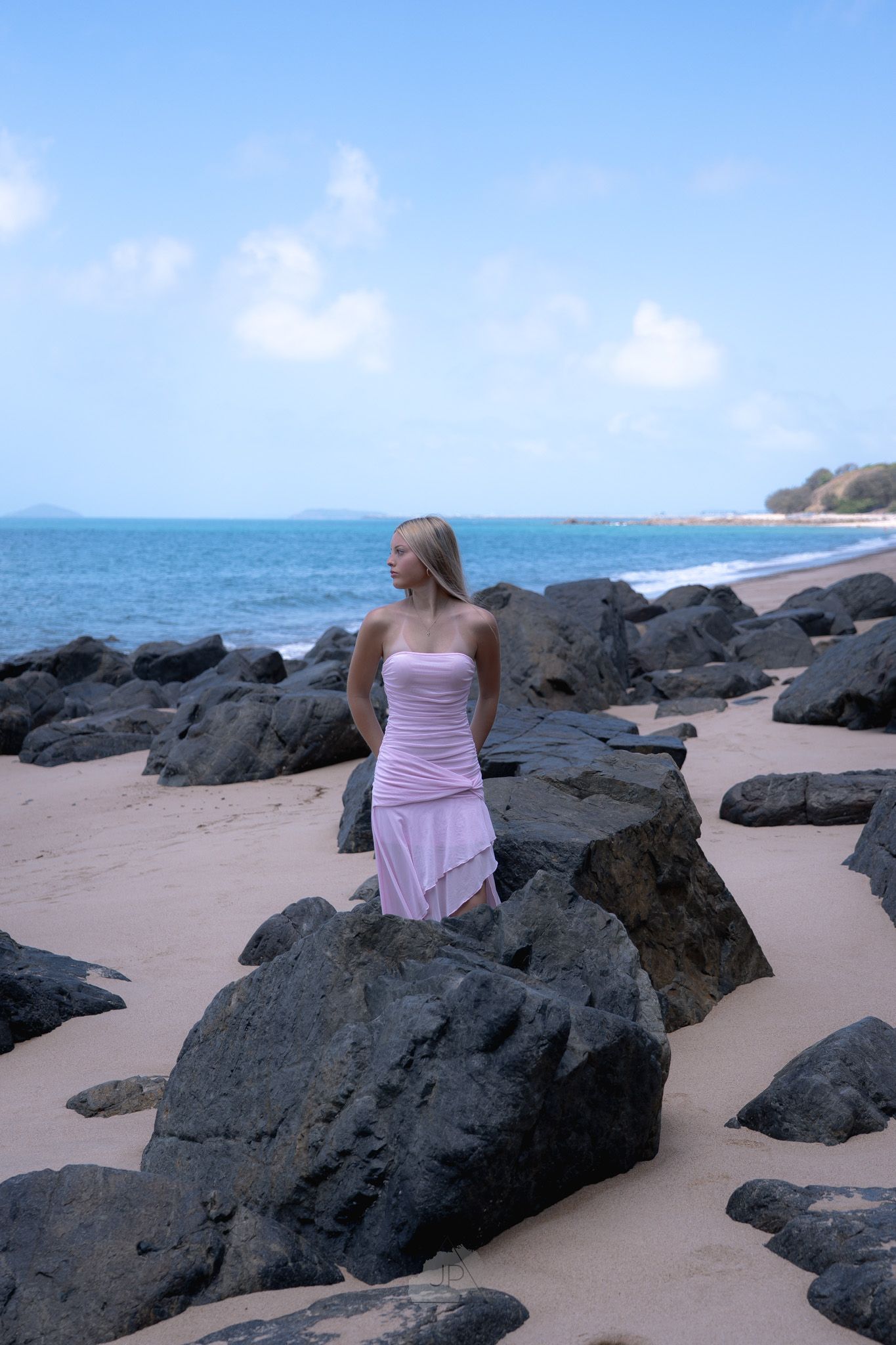 A person in pink dress sits on dark volcanic rocks along a sandy beach with ocean views and blue sky.