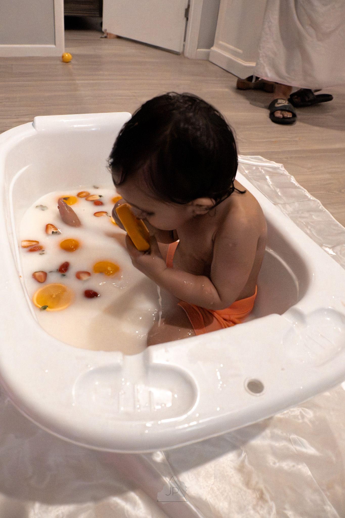 A young child bobbing for fruit slices in a white basin filled with water.
