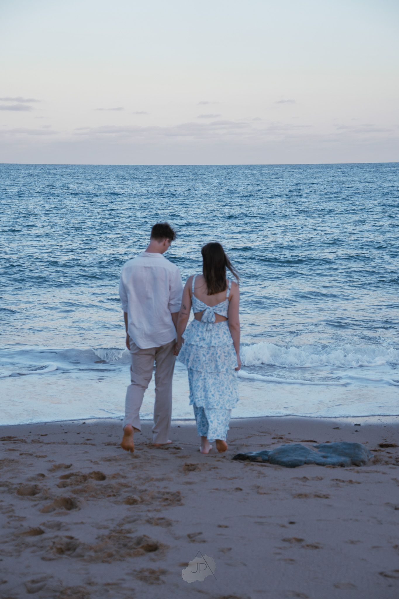 A couple in light colored outfits walks along a serene beach at sunset with gentle waves rolling in.