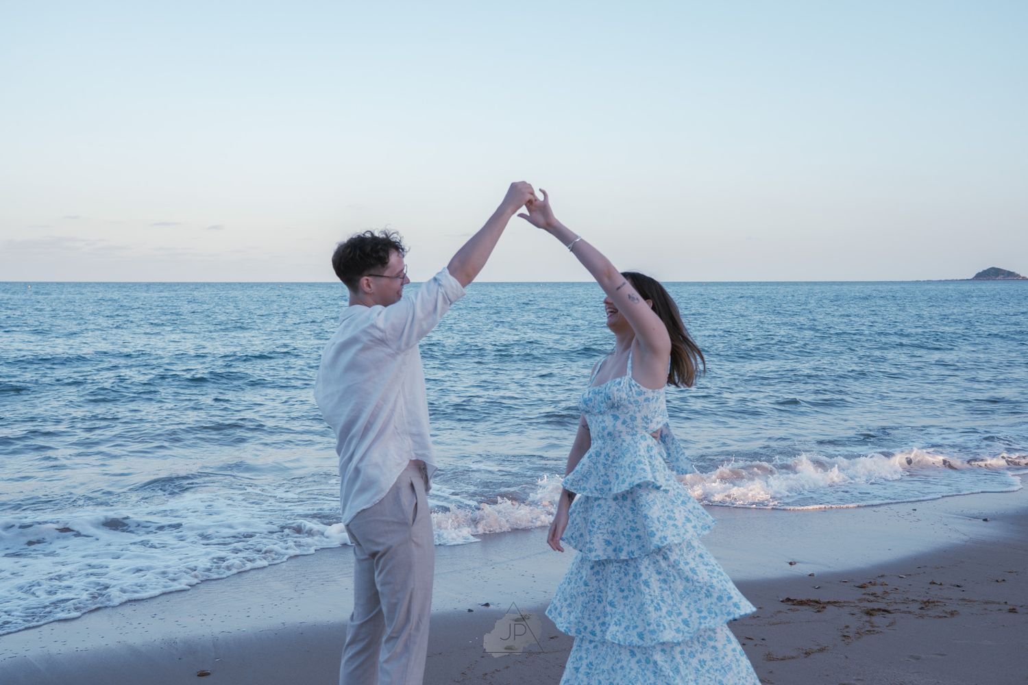 A couple in white and blue outfits walks along the beach during sunset holding hands and raising their arms together.
