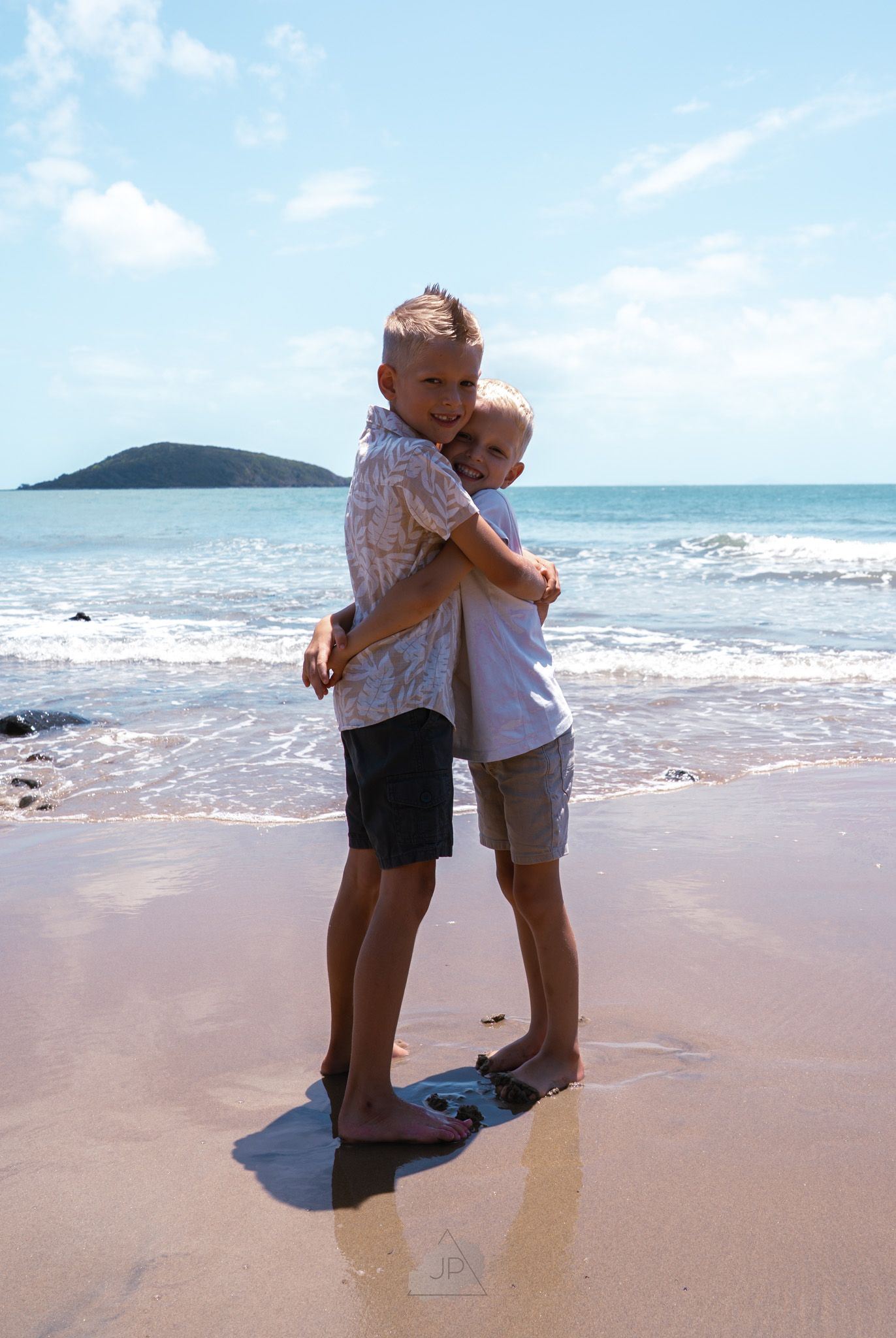 Two people embrace on a sandy beach with ocean waves and a distant island visible in the background.