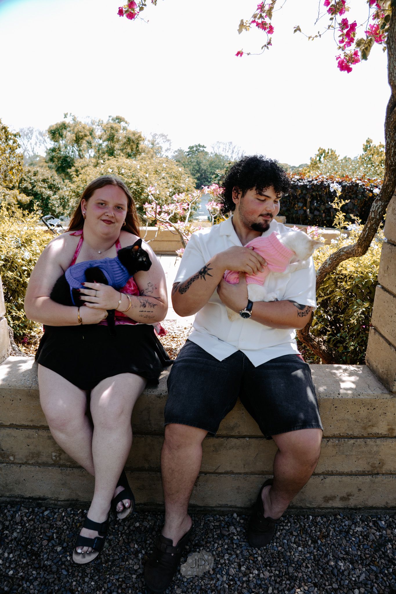 Two people sit together on a stone wall while looking at their mobile phones in an outdoor setting.