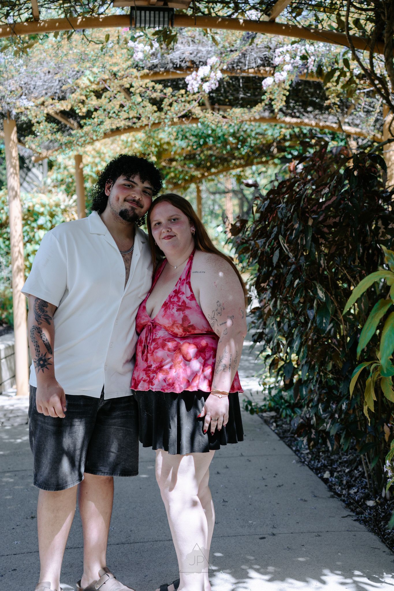 A couple poses together under a shaded walkway in summer attire, with one wearing a pink floral top and black skirt.