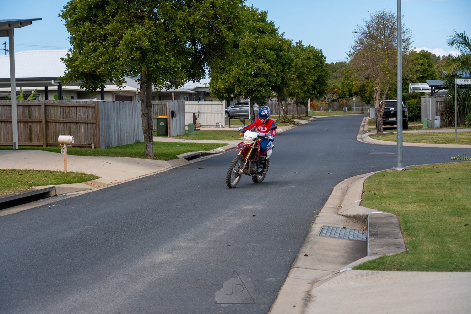 Cyclists ride along a curved suburban street lined with trees and grass on a sunny day.