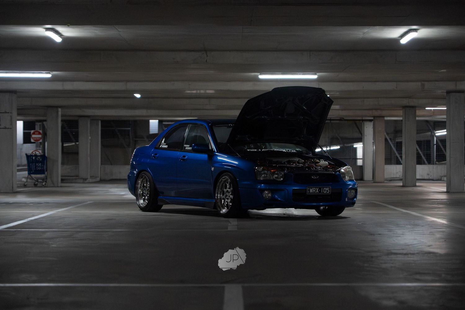 Multiple blue sports cars parked in a dimly lit underground parking garage with fluorescent lighting overhead.