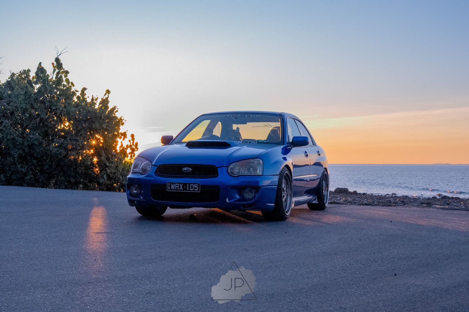 A blue sports car sits on a scenic cliff edge as the sun sets over the horizon.