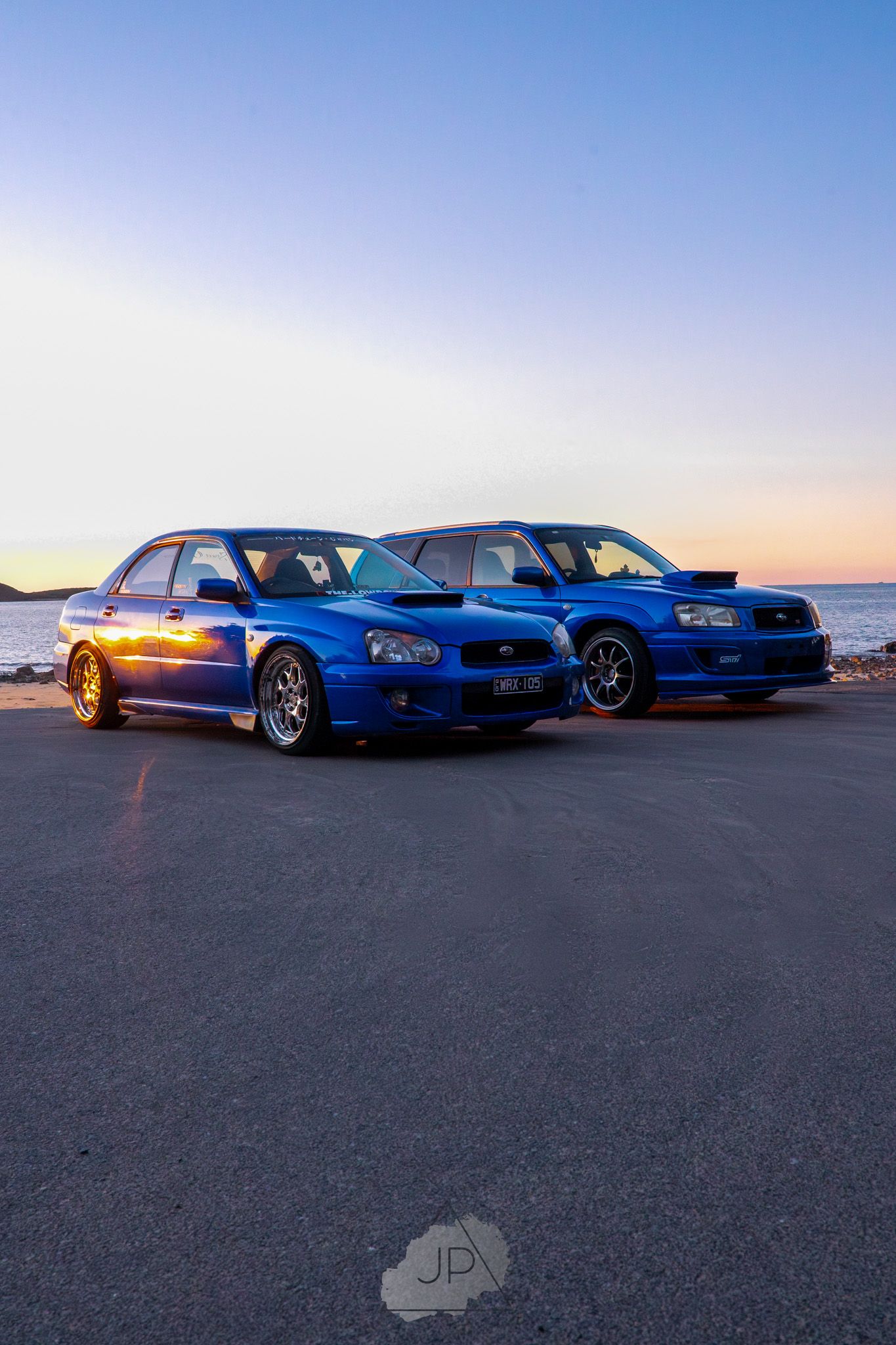 Two blue Subaru WRX STI sports cars parked side by side at sunset against an ocean backdrop.