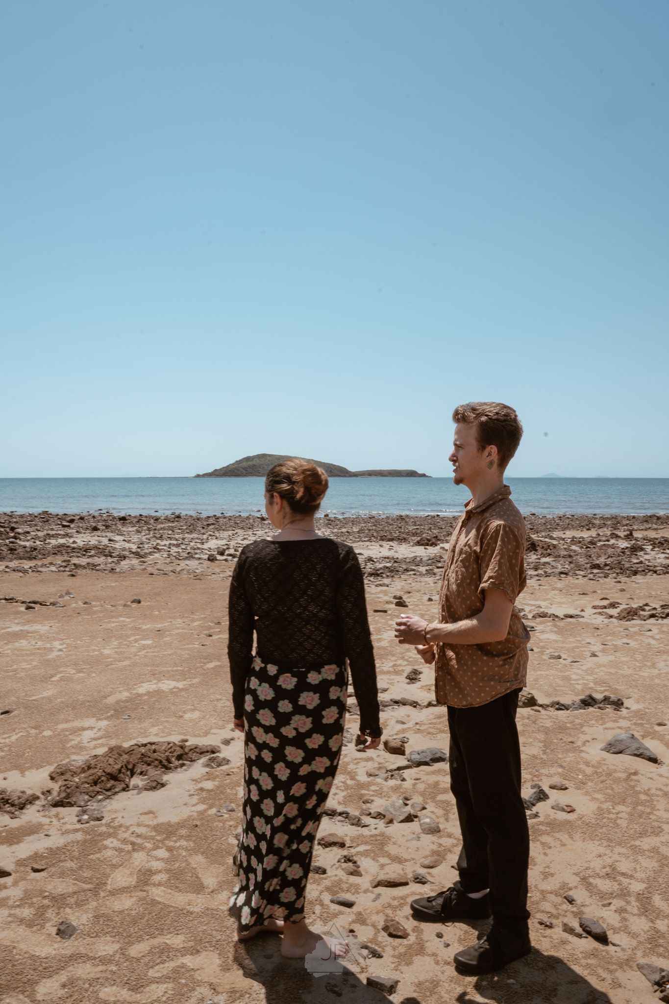 Two people stand on a rocky desert terrain overlooking a distant ocean under bright blue skies.