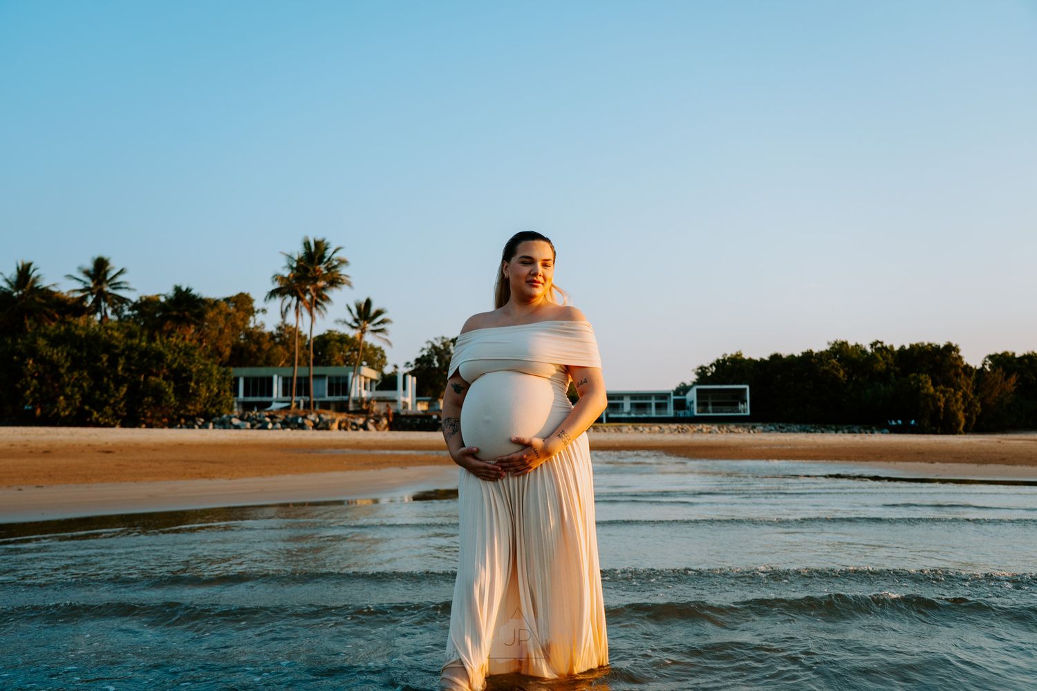 A person in a white dress stands at the beach during sunset while waves gently roll onto the shore.