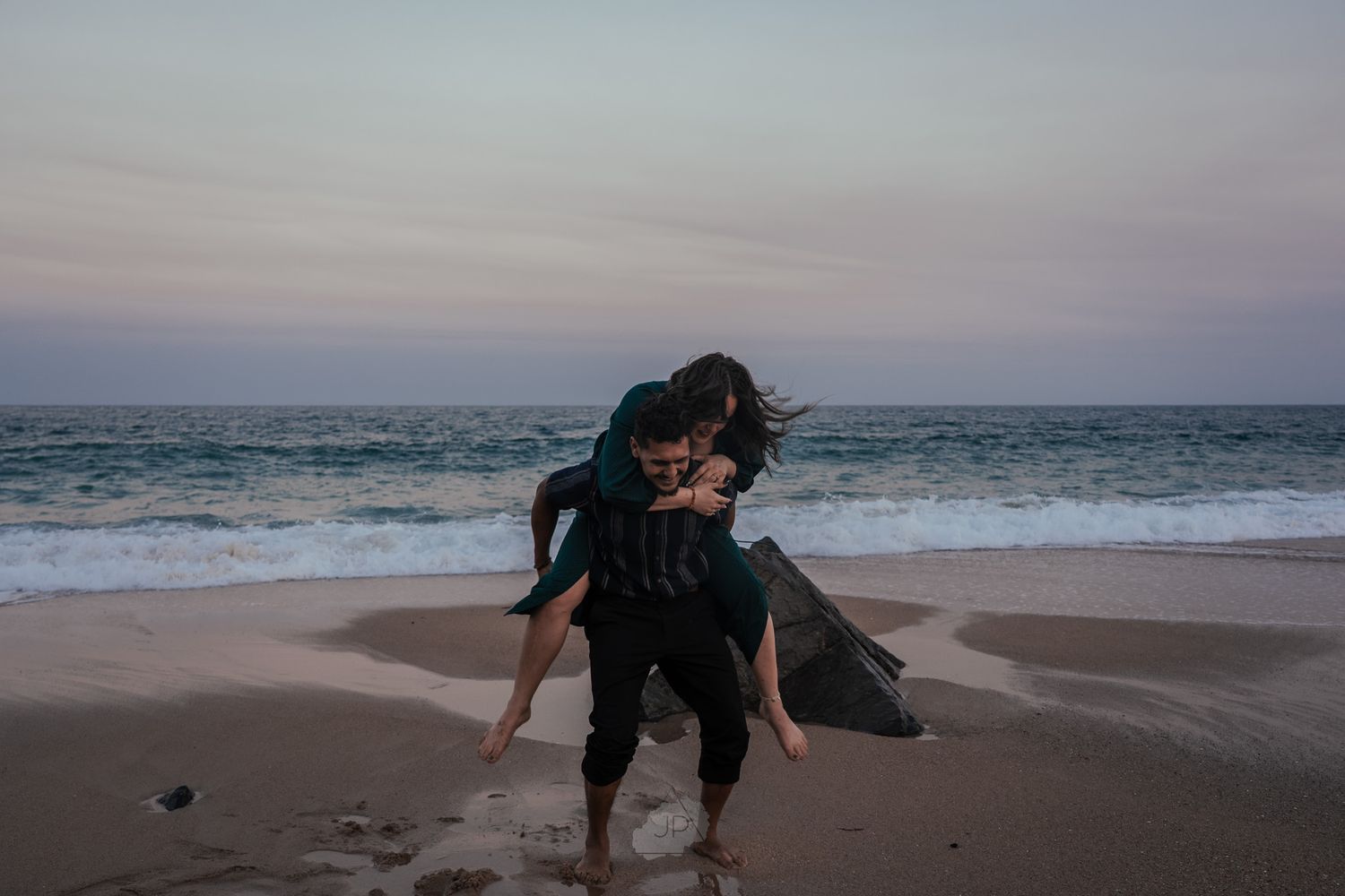 A person in dark clothing walks along a misty beach shoreline at sunset while waves crash against the sand.